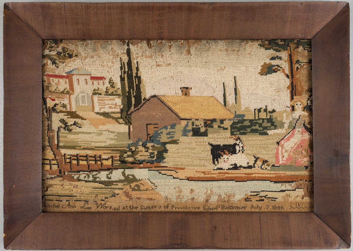 A needlework framed in a brown wooden frame. The needlework depicts a house with a yellow roof and chimney in the center, with trees in the background and bushes in front. There is a bridge on the left with a light blue stream running under it across the image. On the right, there is a light-skinned, blonde haired woman in a pink dress and yellow bonnet. To her left, there is a black goat, a white goat, and a black and tan dog. In the left background, there is a white stone building with a blue and red roof. The bottom of the needlework reads: “Rachel Ann Lee worked at the Sisters of Providence School of Baltimore July 3, 1846.”