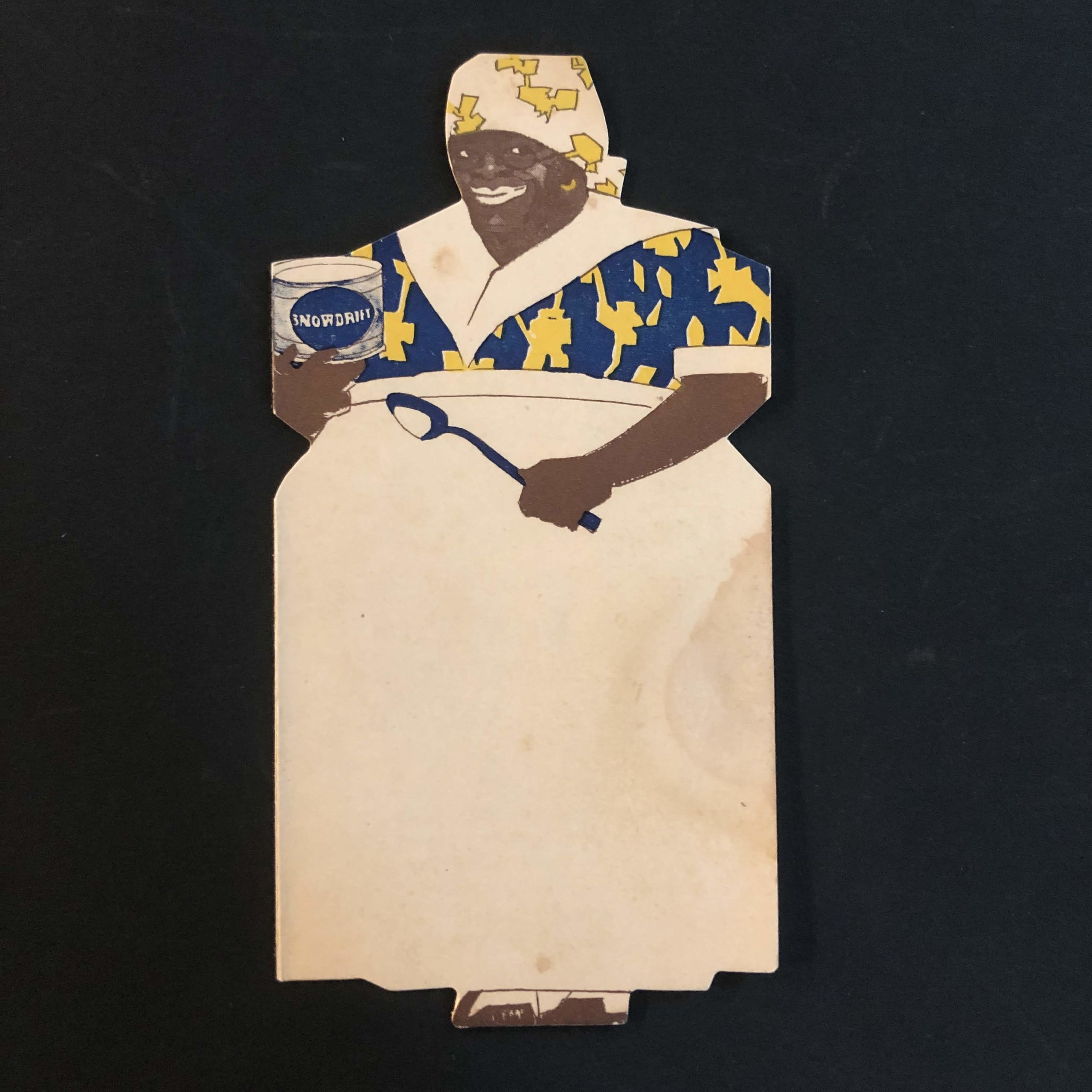 Photograph of a trade catalog in the shape of a dark-skinned woman wearing a kerchief over her head wearing a large white apron over a print dress holding a spoon and a can of "Snowdrift" shortening.
