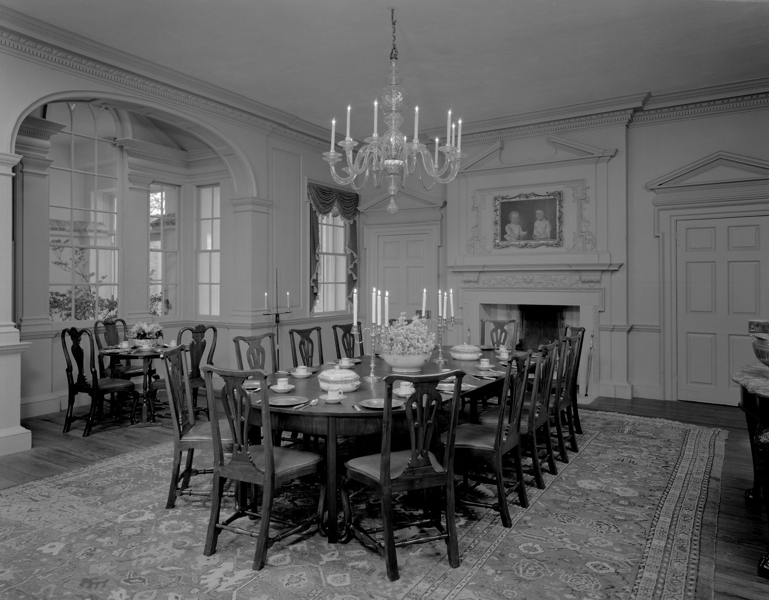 Black and white photograph of a finely furnished dining room with carpet, chandelier, and a large table at center.