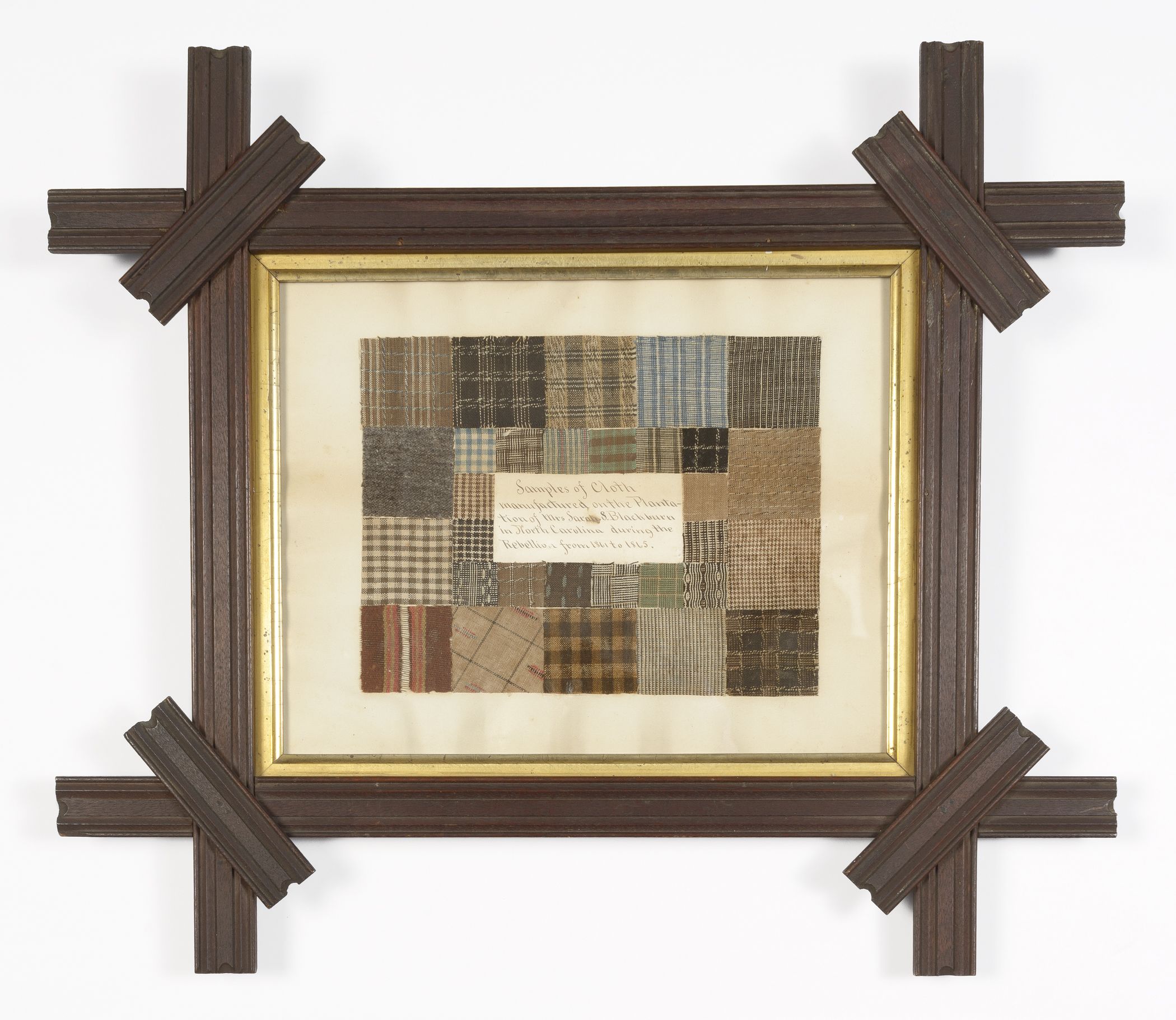 A framed collection of plaid fabric squares in a variety of colors, arranged in a rectangular shape with a rectangular piece of paper in the center. The paper reads: “Samples of Cloth manufactured on the Plantation of Mrs. Sarah S. Blackburn in North Carolina during the Rebellion from 1861 to 1865.” The frame is made of brown wood.