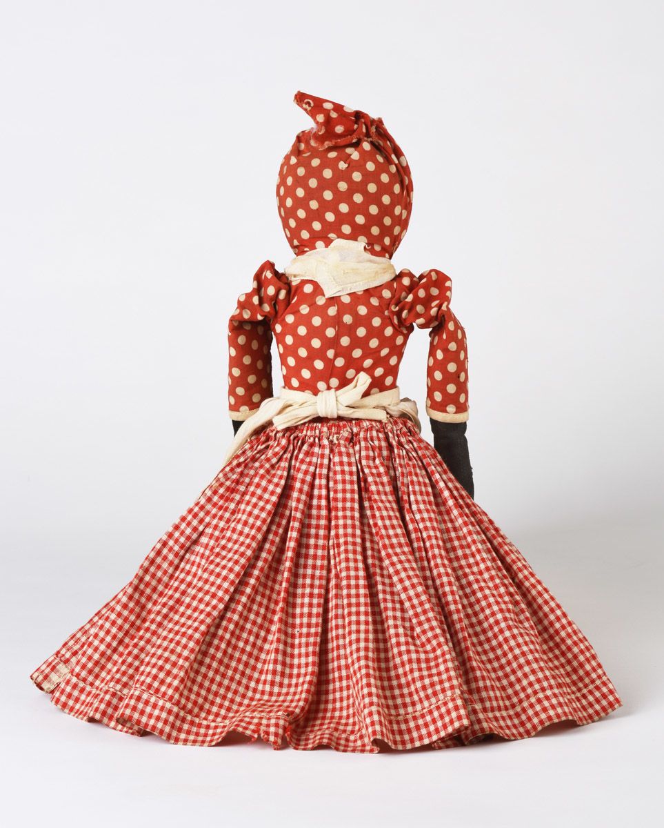 Back of a two-sided cloth doll with a skirt that flips from one side to the other to reveal the torso of a dark-skinned female figure with a red and white dress. Her bodice is red and white polka-dot, her skirt is red and white gingham. She is wearing a simple white apron and a red and white polka-dot headwrap.
