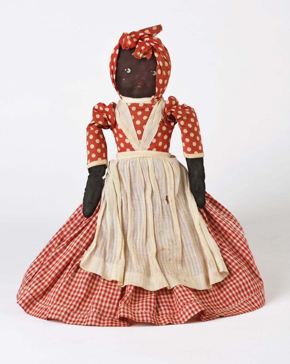 Front of a two-sided cloth doll with a skirt that flips from one side to the other to reveal the torso of a dark-skinned female figure with a red and white dress. Her bodice is red and white polka-dot, her skirt is red and white gingham. She is wearing a simple white apron and a red and white polka-dot headwrap.