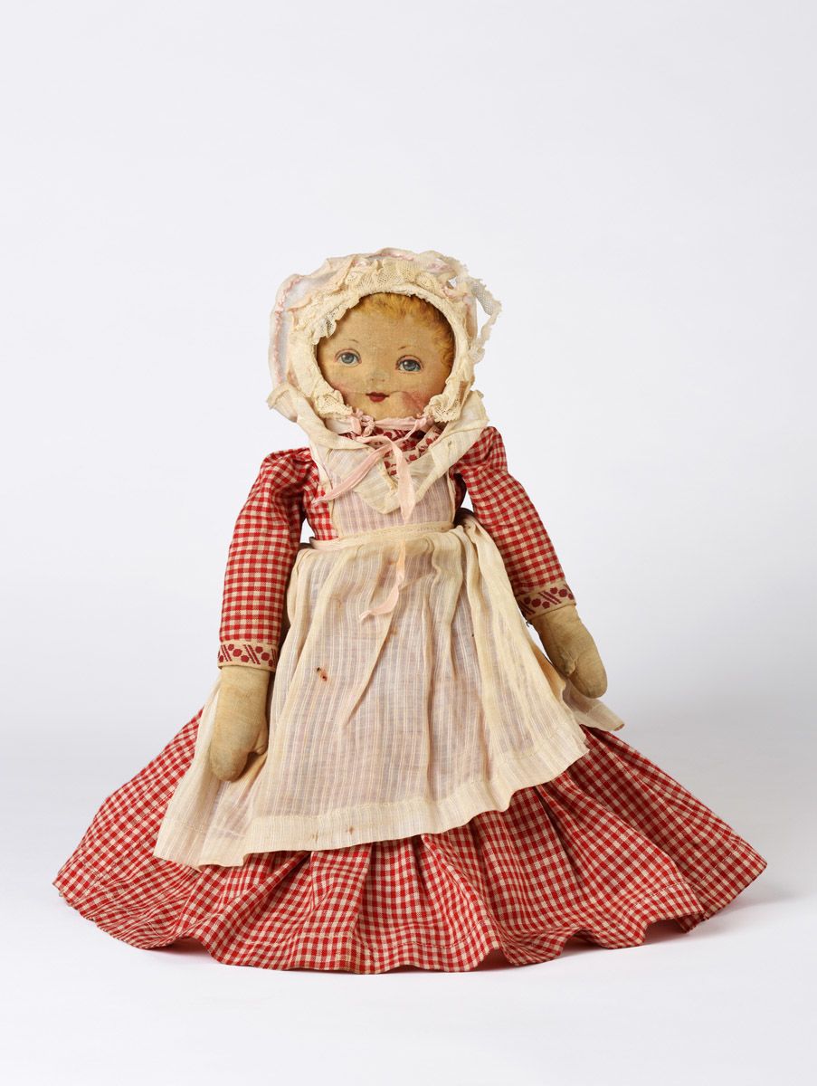 Front of two-sided cloth doll with a skirt that flips from one side to the other to reveal the torso of a female figure with light skin, blue eyes, and blonde hair. She is dressed in a red and white gingham dress with trim. She is wearing a white ruffled apron and a white cap with lace. She has painted facial features.