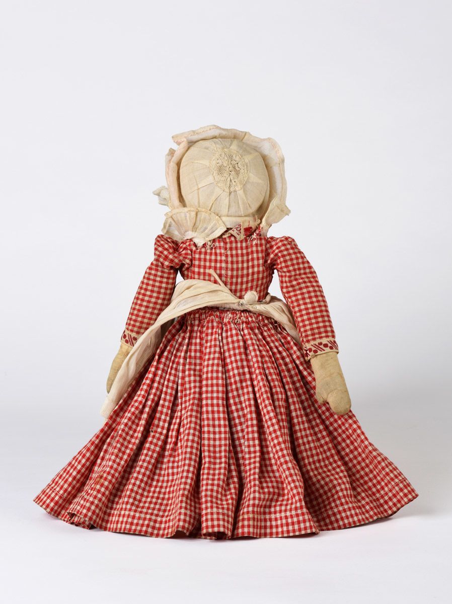 Back of two-sided cloth doll with a skirt that flips from one side to the other to reveal the torso of a female figure with light skin, blue eyes, and blonde hair. She is dressed in a red and white gingham dress with trim. She is wearing a white ruffled apron and a white cap with lace. She has painted facial features.